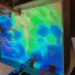 Mit den Händen konnte man eine Sandlandschaft formen. Die sich dabei verändernden Farben stellen unterschiedliche Höhenbereiche dar. Auch eine Wasseroberfläche wird angezeigt. Mit den Händen konnte man eine Sandlandschaft formen. Die sich dabei verändernden Farben stellen unterschiedliche Höhenbereiche dar. Auch eine Wasseroberfläche wird angezeigt.